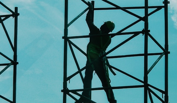 A construction worker wearing a hard hat stands on scaffolding, overseeing the building process below.