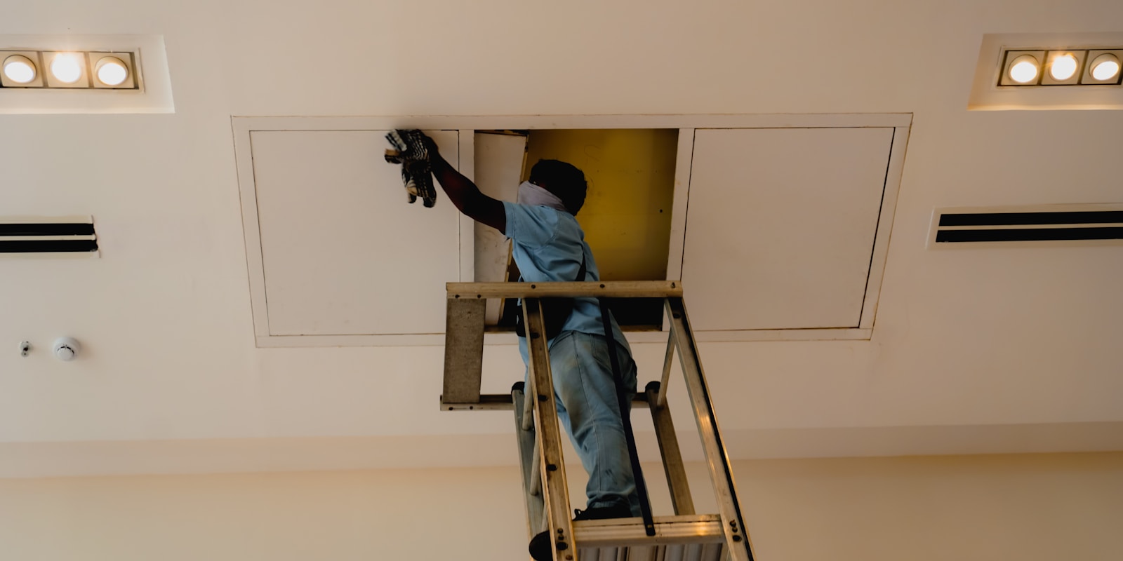 A man on a ladder cleaning a ceiling with a cloth, focused on removing dust and cobwebs.