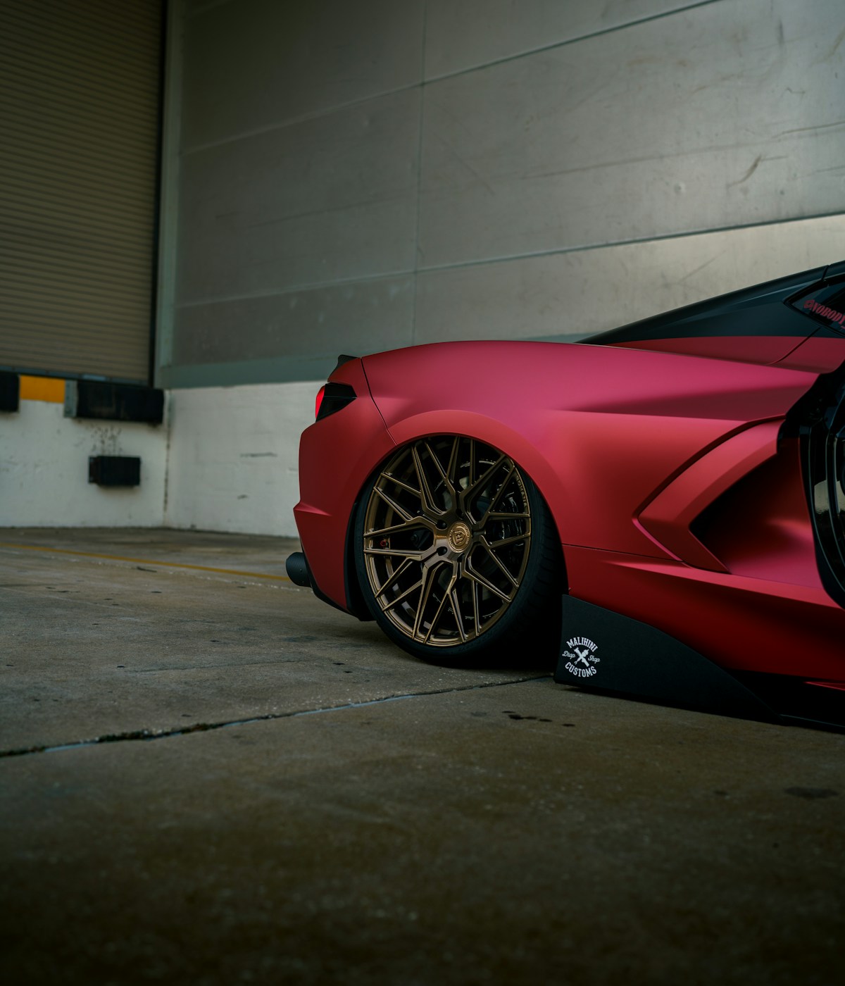 Close-up of the rear section of a matte red luxury sports car with gold wheels, parked in an industrial setting, exuding elegance and power.