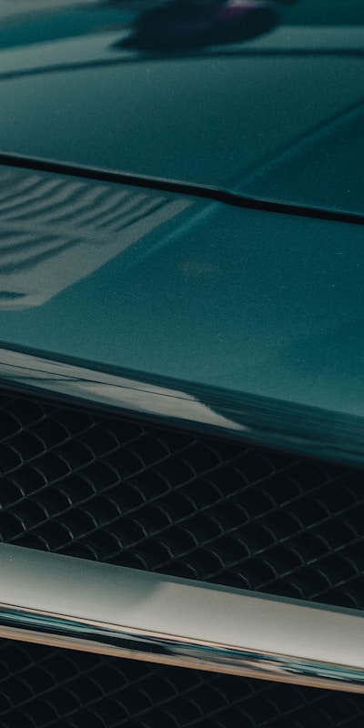 Close-up of a sleek, glossy car hood in dark teal, featuring a subtle reflection and a black mesh grille below, conveying elegance and luxury.