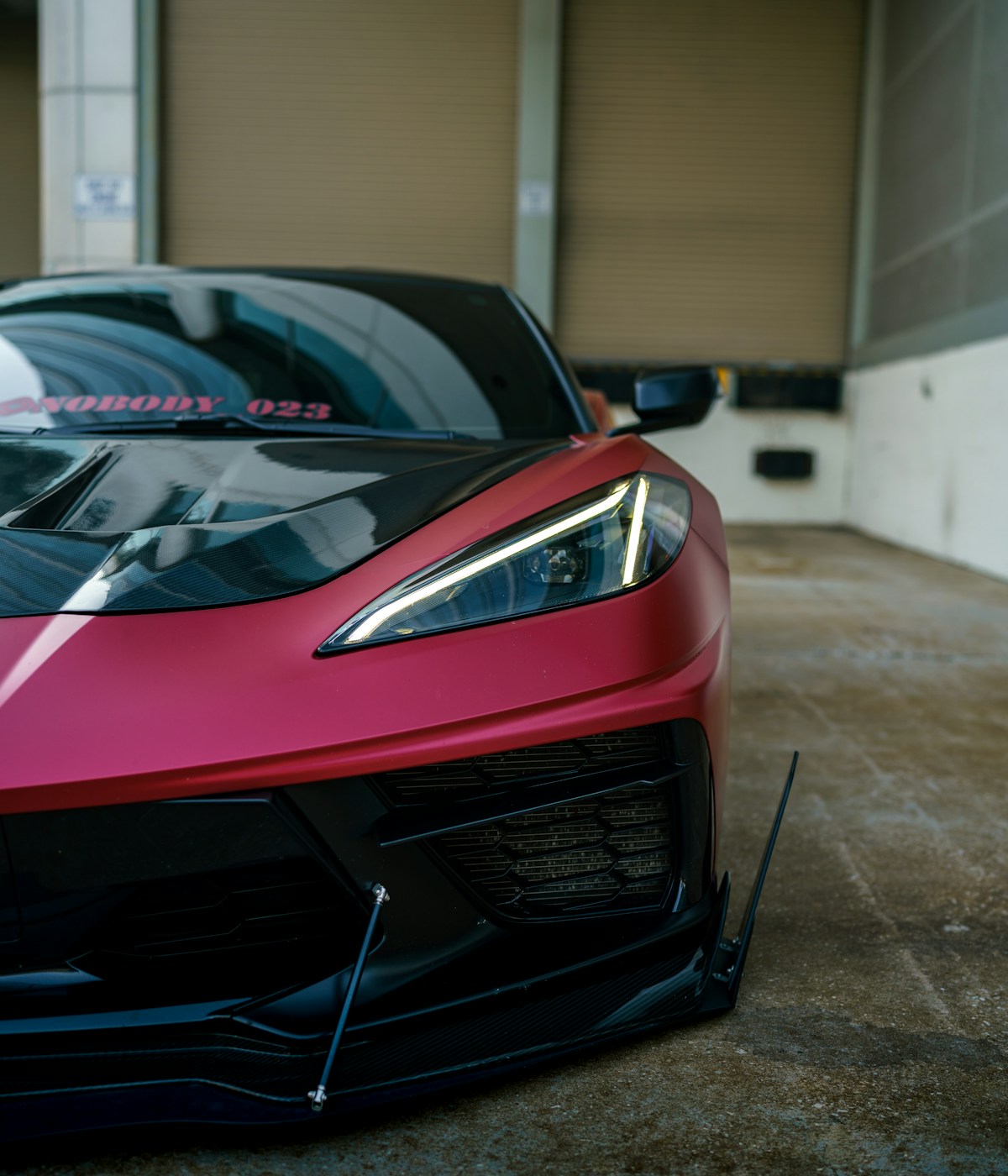 Close-up of a sleek red sports car with a black carbon fiber hood, parked in an industrial setting. The car's sharp headlight and aerodynamic design convey speed and luxury.