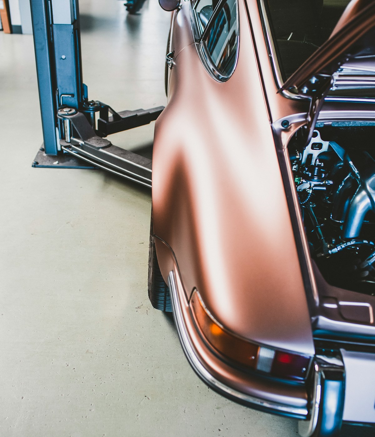 Close-up of a shiny, copper-colored classic car with its engine exposed, positioned on a hydraulic lift in a garage with a clean floor.