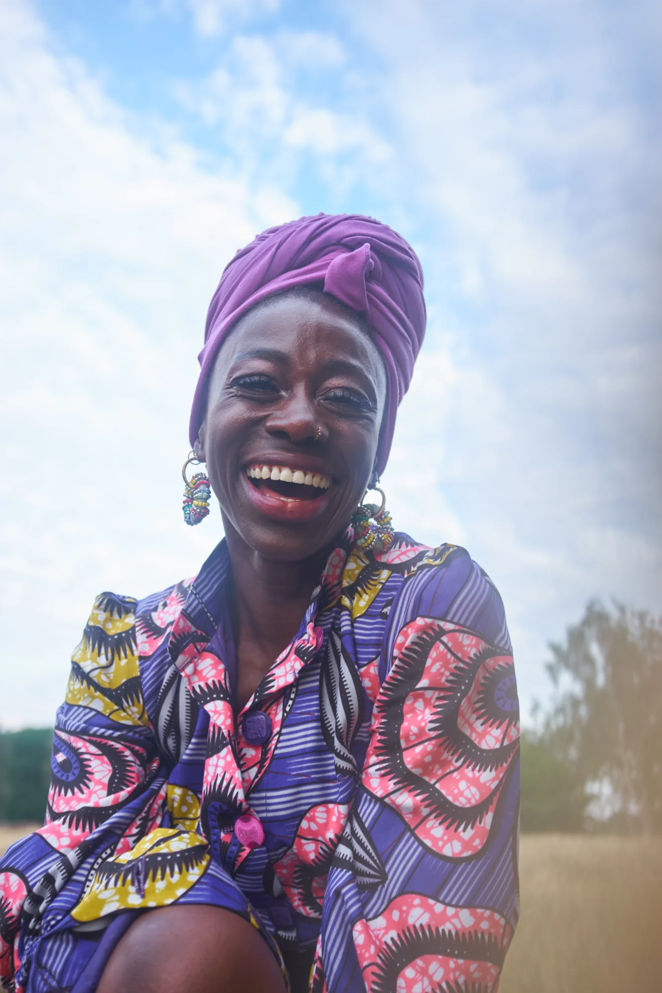 A woman smiling brightly in a colourful patterned outfit and headwrap against a blue sky