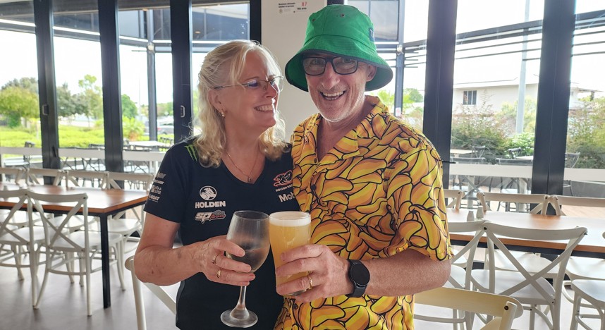 A smiling beer buff and buffette enjoying drinks at a modern bar, with the man wearing a bright yellow banana-print shirt and green bucket hat and the woman holding a wine glass, both laughing together.